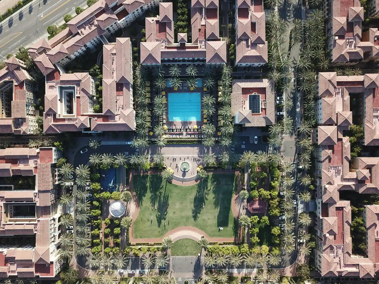 Aerial view of an apartment complex with red tile roofs, palm trees, a swimming pool, courtyard, and landscaped walkways.