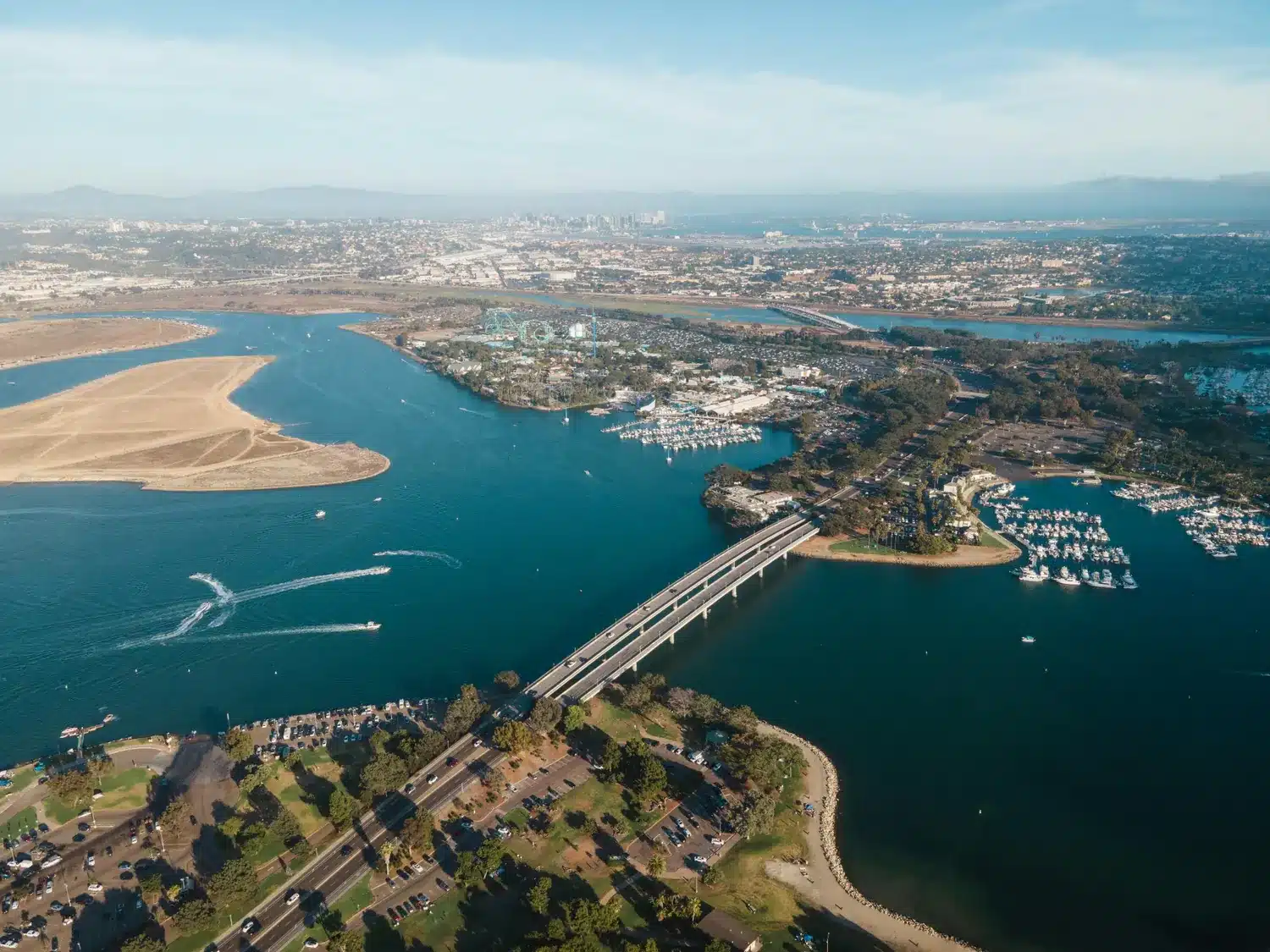 Aerial view of a bay with a bridge, marina, boats, shoreline parks, and city buildings in the distance.