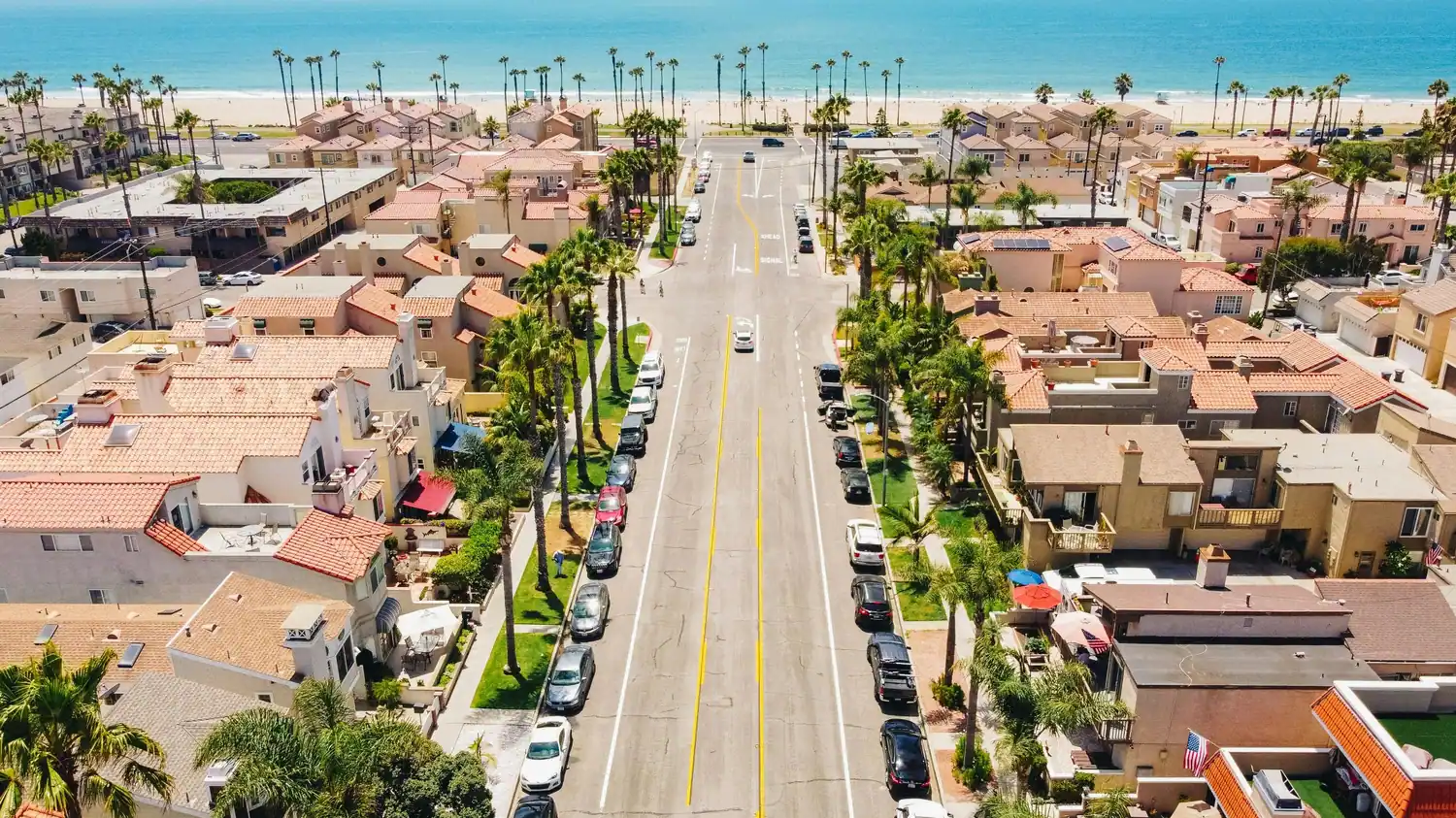 Aerial view of a beachside street lined with homes, palm trees, parked cars, and the ocean in the background.