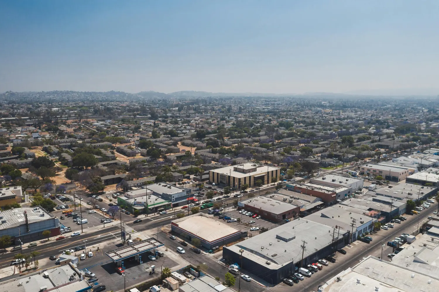 Aerial view of a city neighborhood with commercial buildings, streets, parking lots, homes, and hazy hills in the distance.