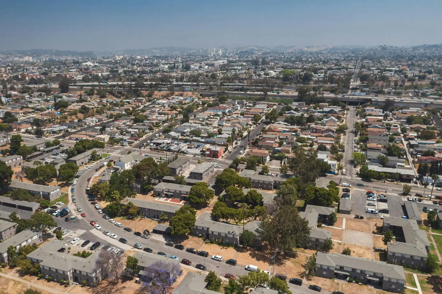 Aerial view of a city neighborhood with homes, apartment buildings, streets, trees, and a freeway in the distance.