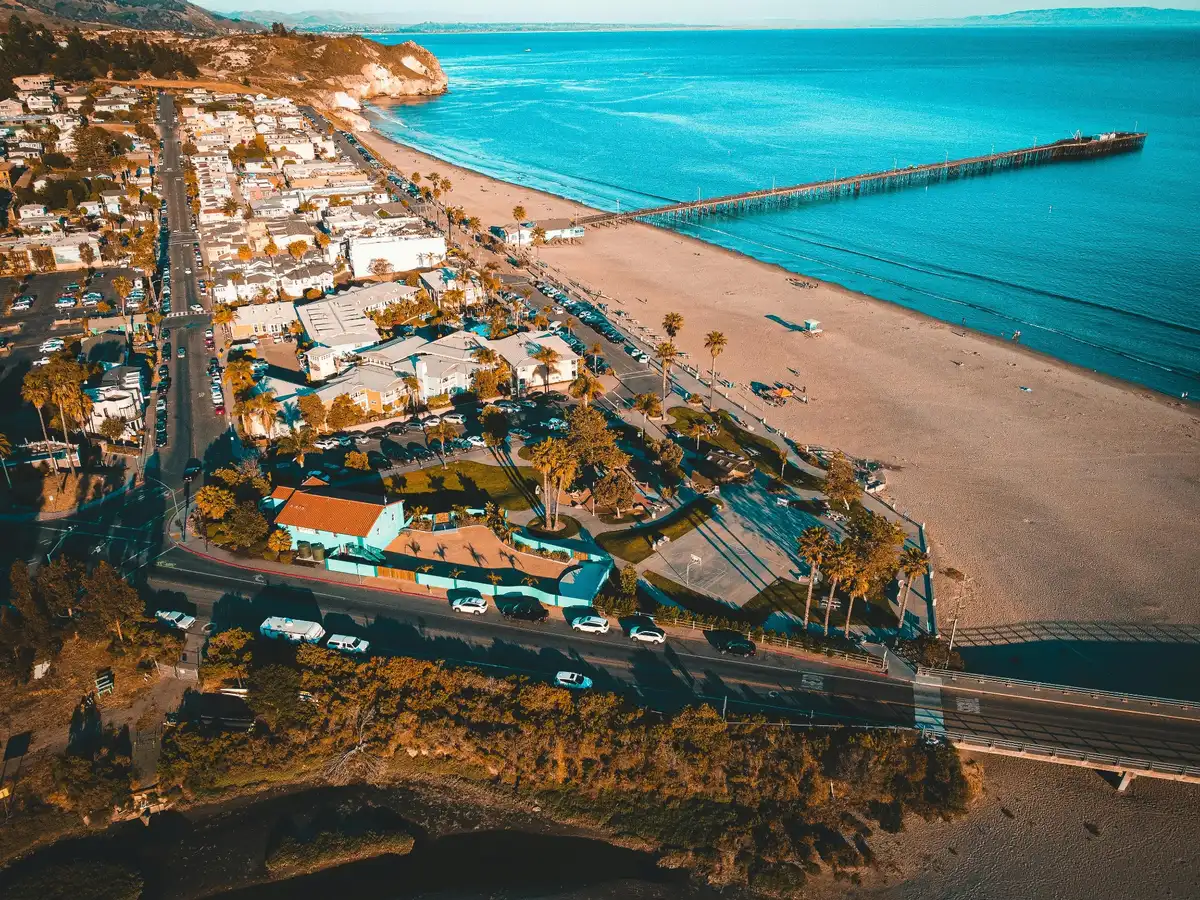 Aerial view of a coastal beach community with a long pier, shoreline homes, palm trees, blue ocean, and nearby cliffs.