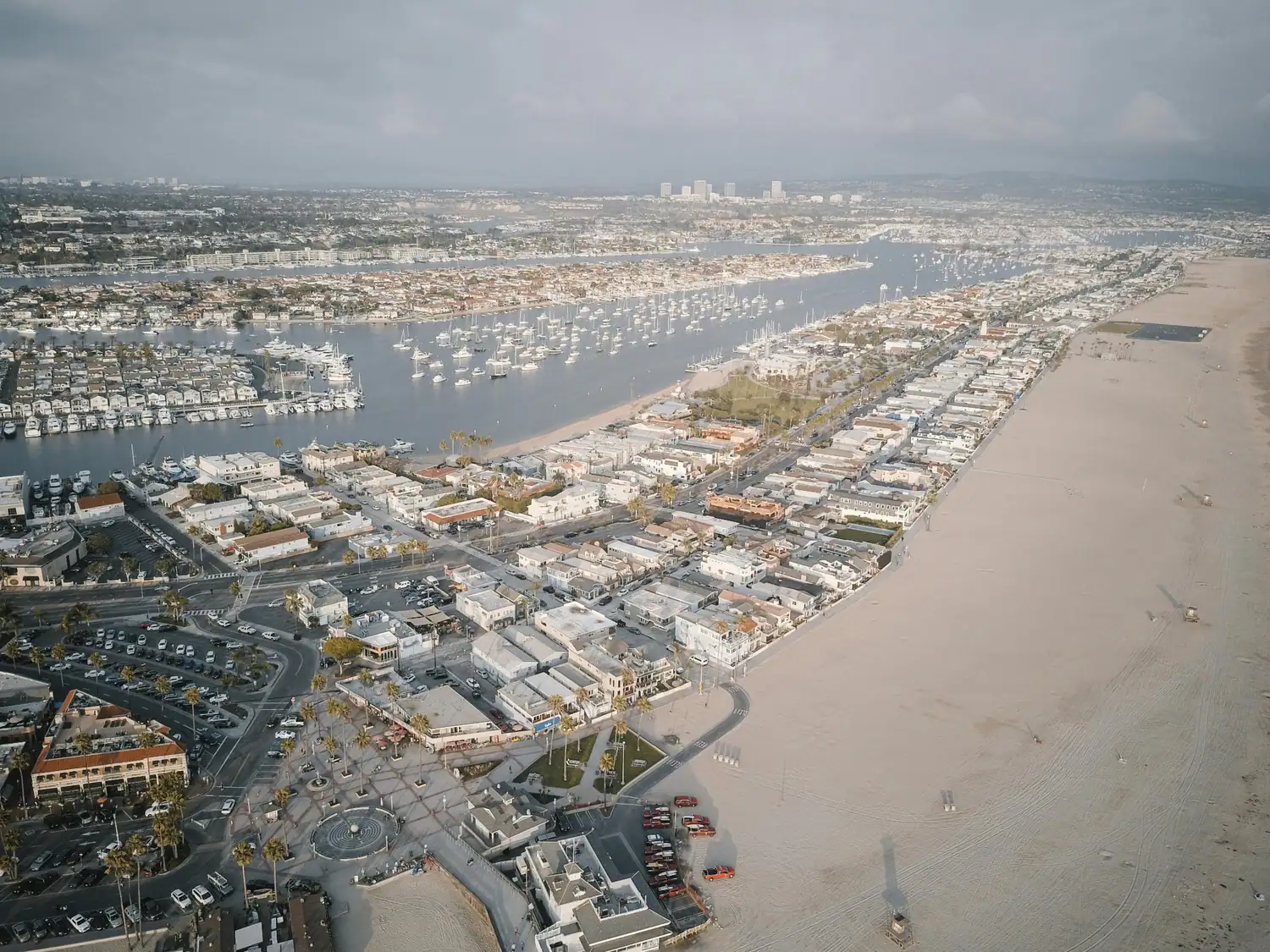 Aerial view of a coastal harbor with boats, beachfront homes, a marina, sandy shoreline, and city buildings in the distance.