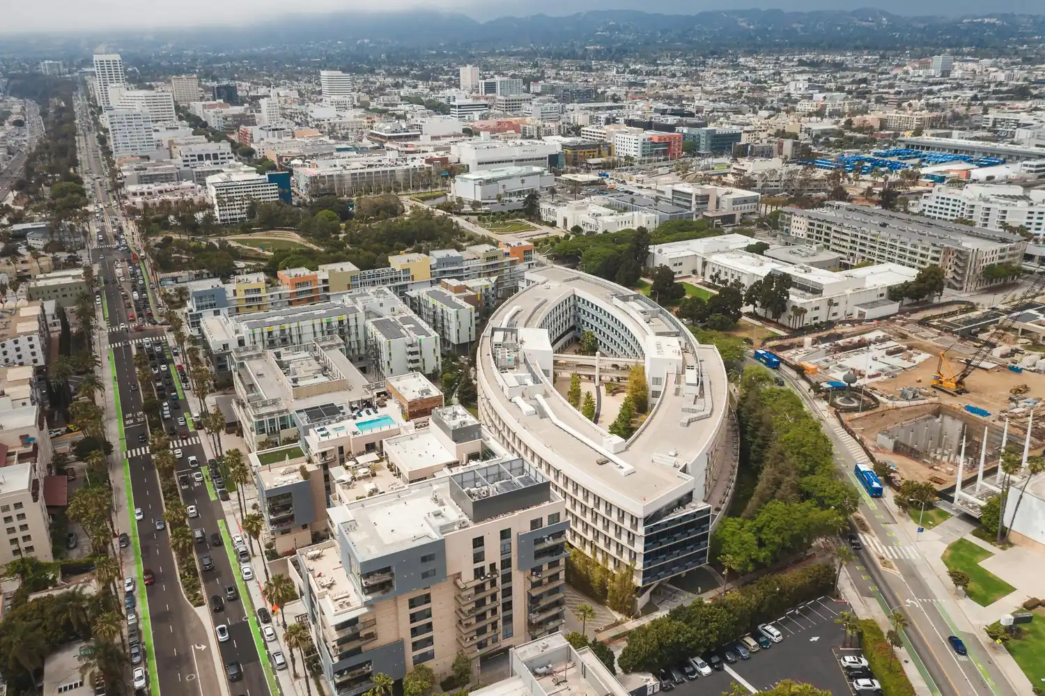 Aerial view of a downtown area with modern buildings, busy streets, parking areas, trees, and hills in the background.