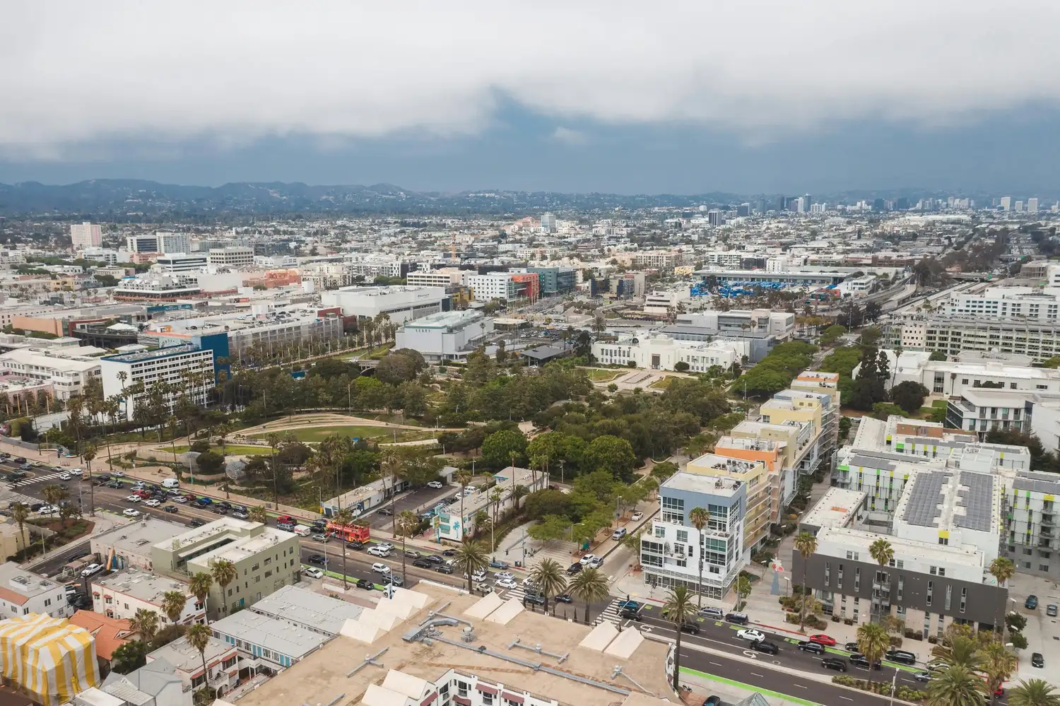 Aerial view of a downtown area with modern buildings, streets, parks, trees, and mountains under a cloudy sky.