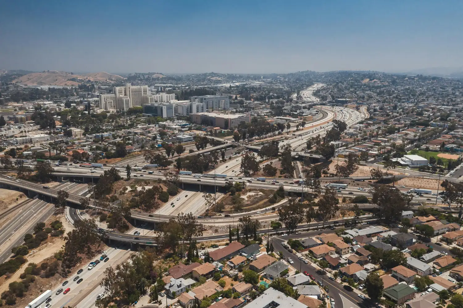 Aerial view of a city freeway interchange with nearby neighborhoods, buildings, traffic, and hills in the distance.