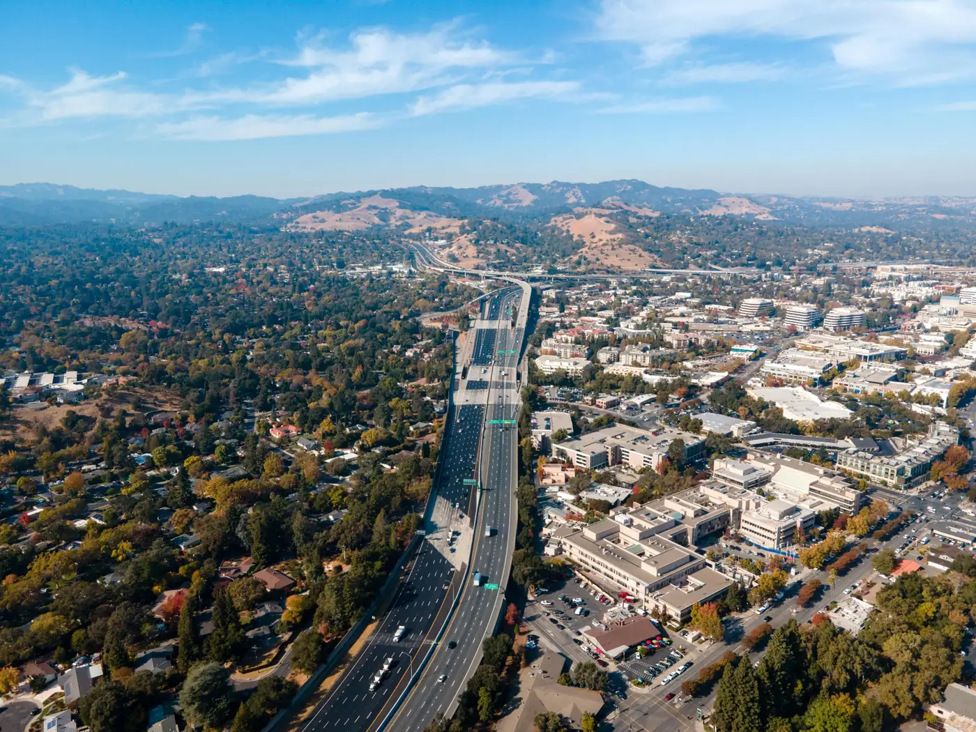 Aerial view of a freeway running through a city with homes, commercial buildings, trees, and rolling hills in the distance.