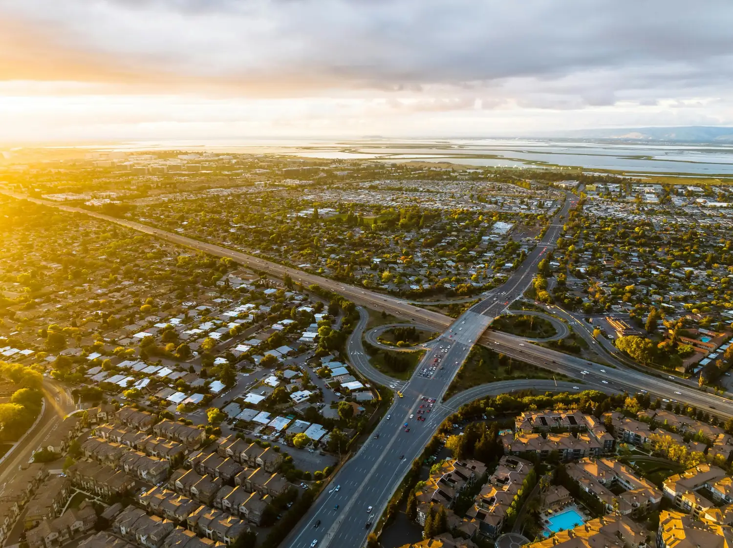 Aerial sunset view of a freeway interchange running through a city with neighborhoods, trees, and water in the distance.
