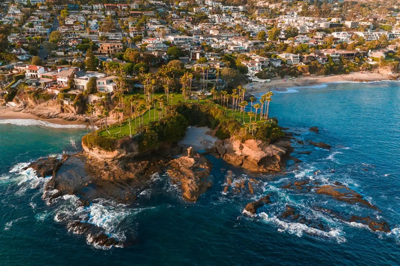 Aerial view of a coastal neighborhood with oceanfront homes, rocky shoreline, palm trees, and blue water.