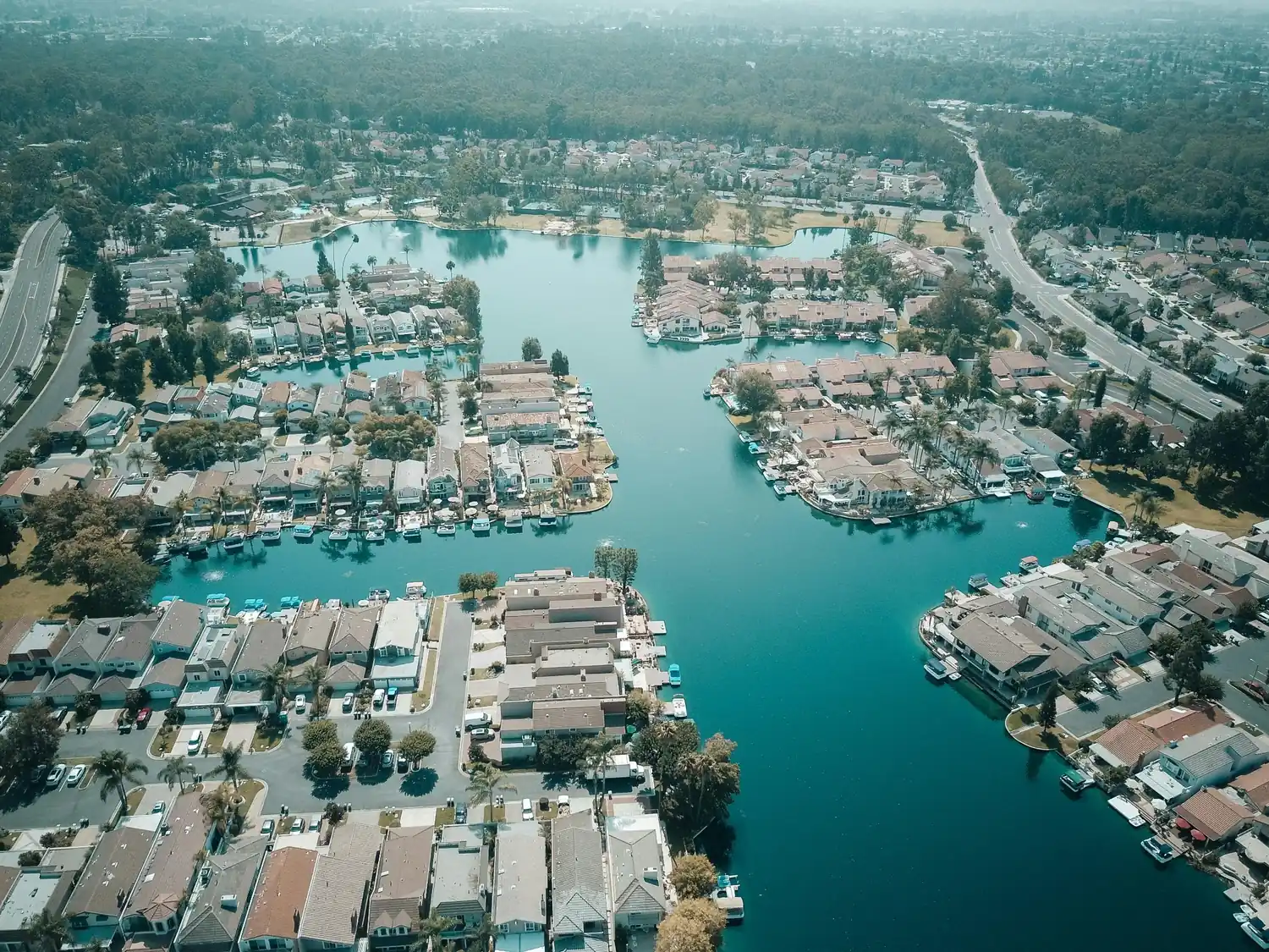 Aerial view of lakeside homes with docks, calm blue water, nearby roads, trees, and surrounding neighborhoods.