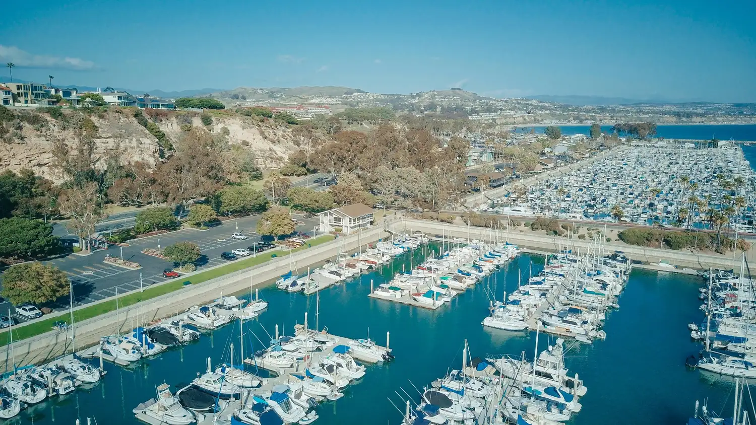 Aerial view of a coastal marina with boats, nearby cliffs, parking areas, roads, and blue ocean water.