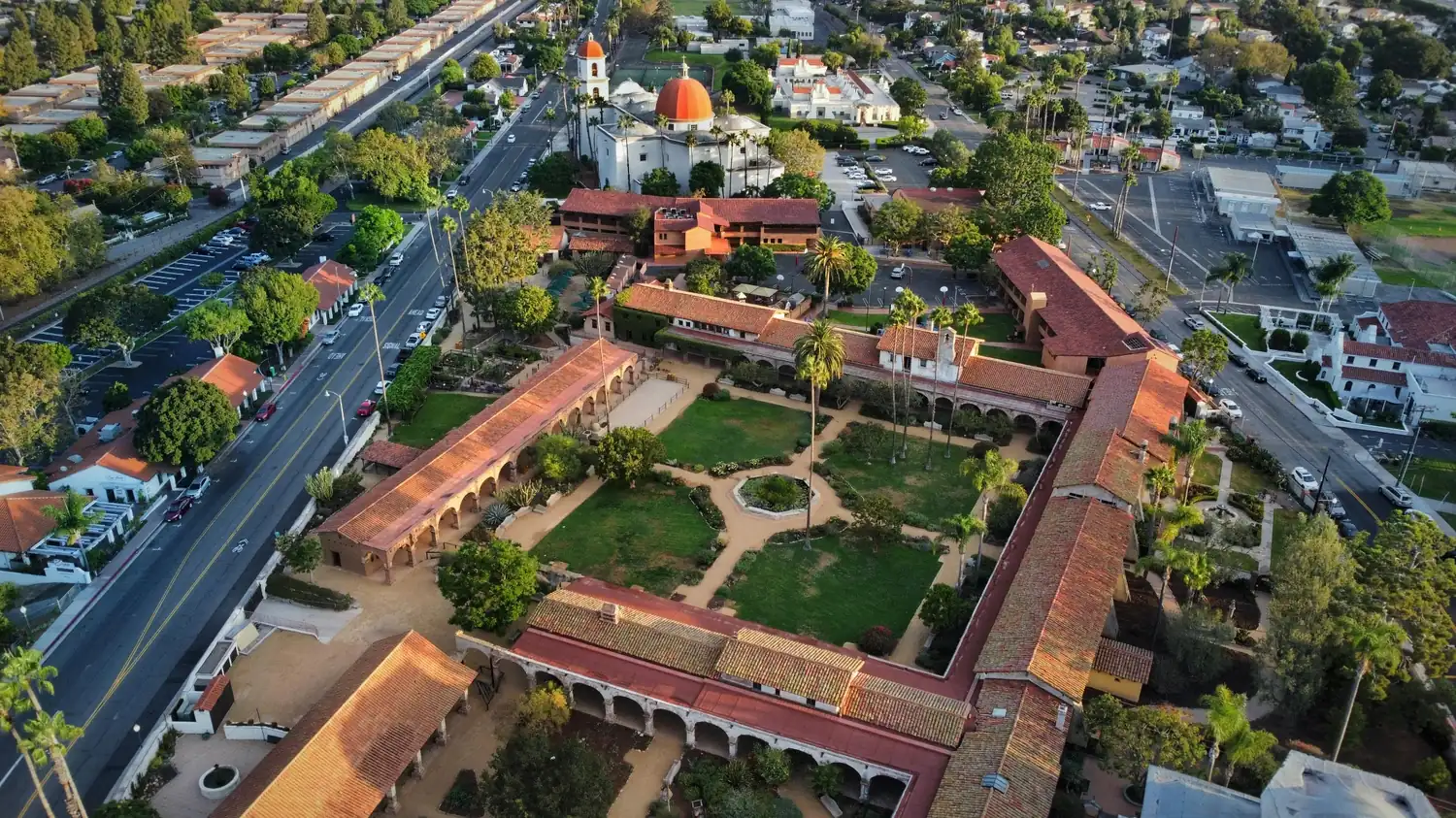 Aerial view of a historic mission-style courtyard with red tile roofs, gardens, palm trees, nearby streets, and a domed church.