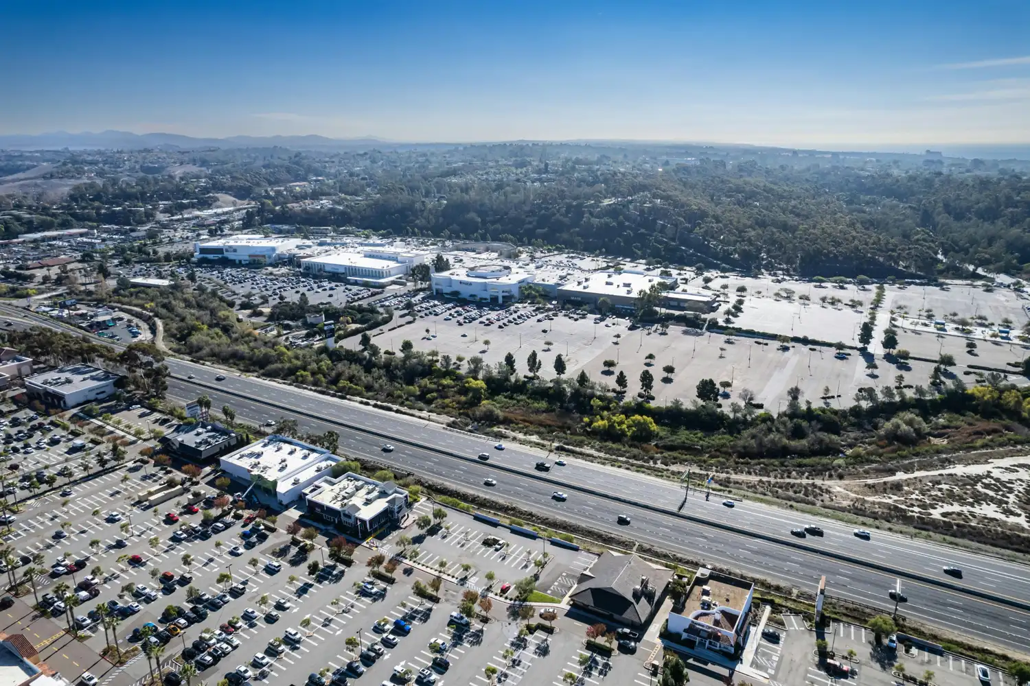 Aerial view of a shopping center and large parking lots beside a freeway, with hills, trees, and the ocean in the distance.