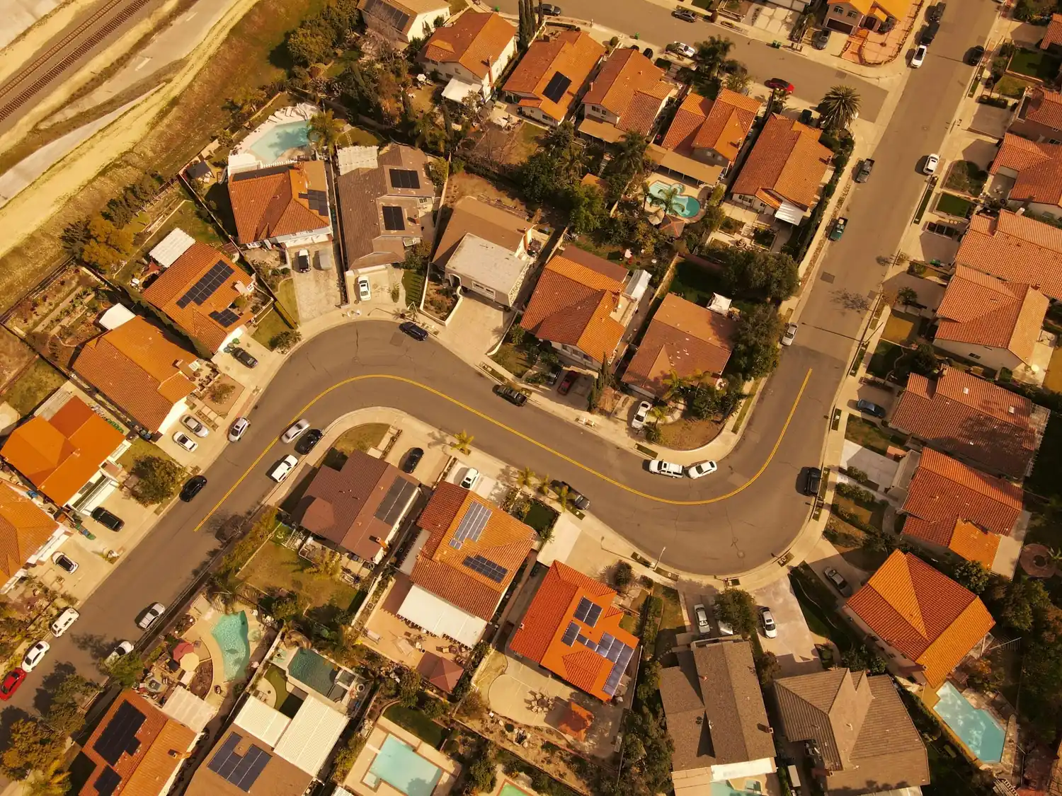 Top-down aerial view of suburban homes with red tile roofs, curved streets, pools, solar panels, and warm sunlight.