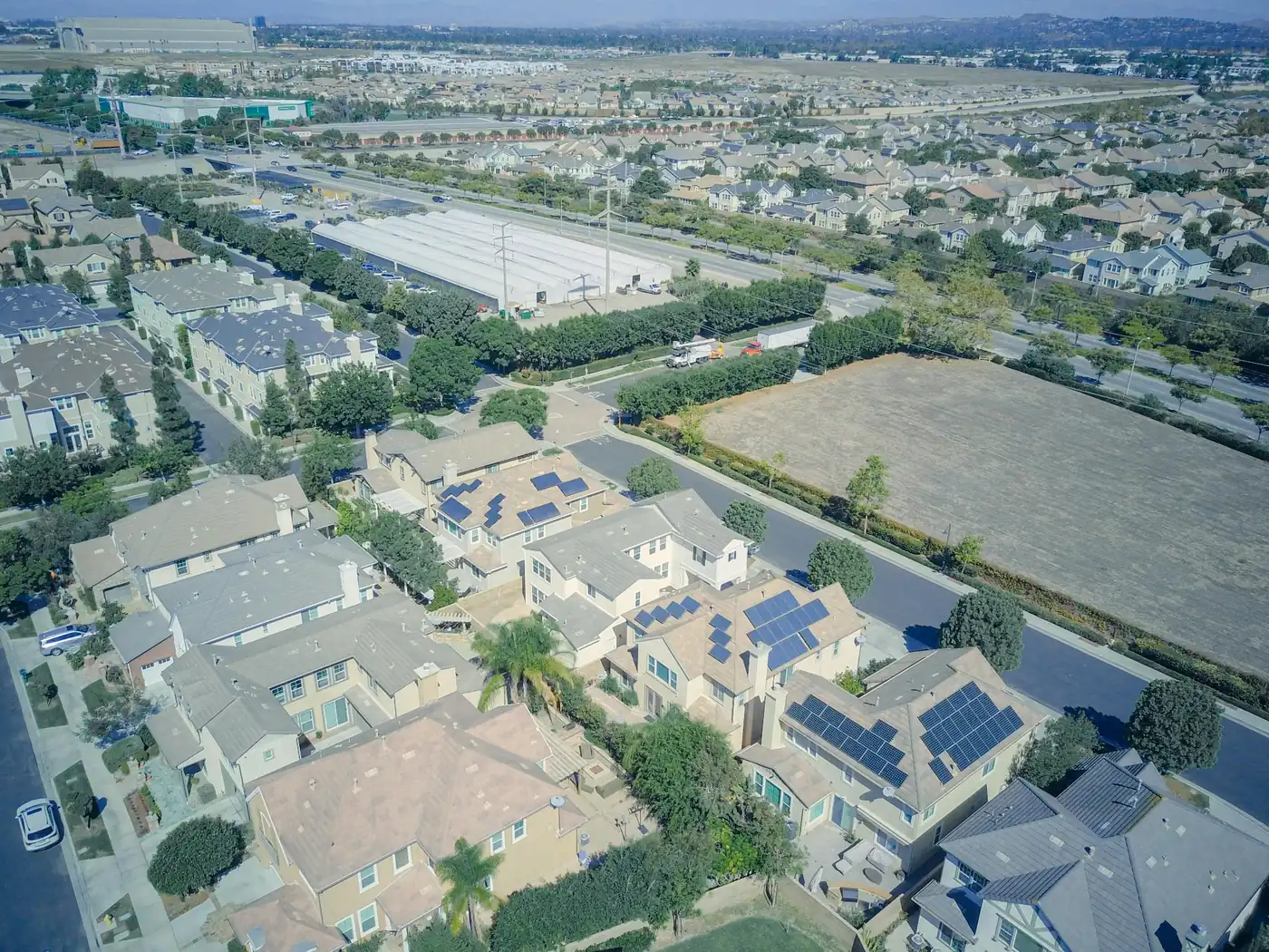 Aerial view of a suburban neighborhood with homes, solar panels, streets, trees, nearby fields, and distant hills.
