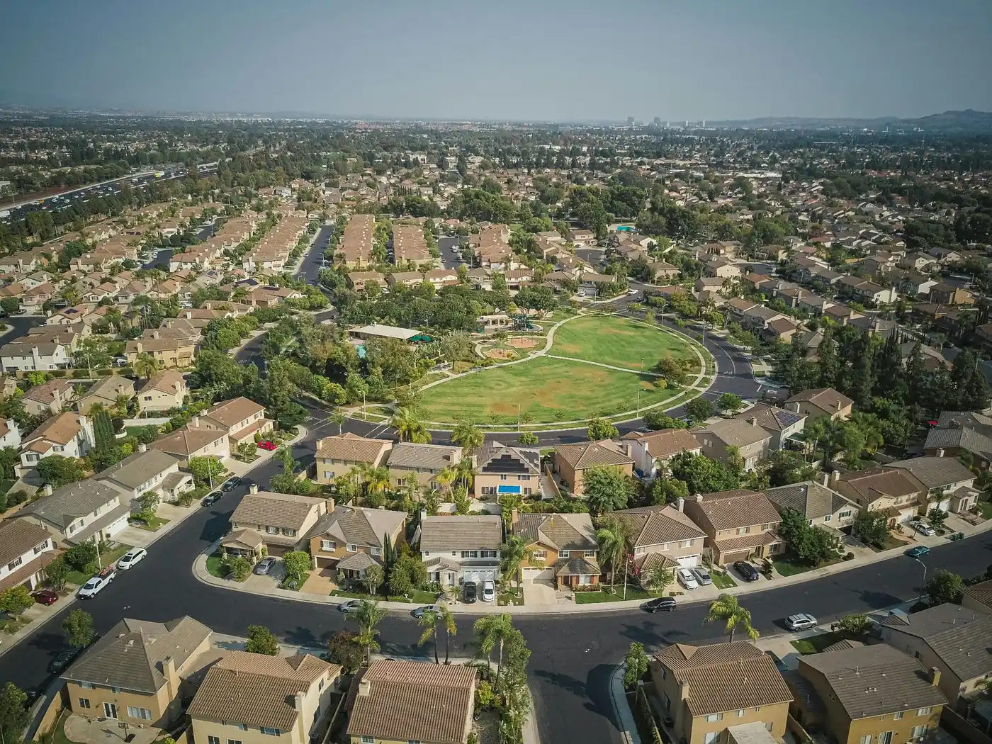 Aerial view of a suburban neighborhood with homes, curved streets, trees, and a large green park in the center.