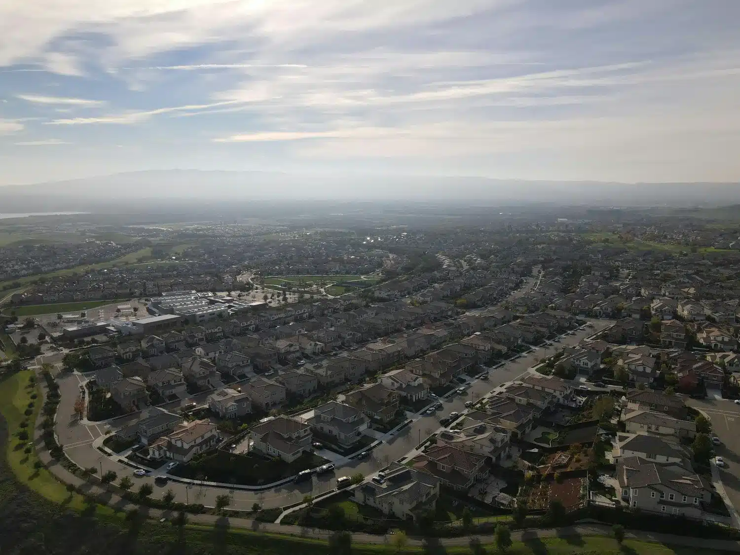 Aerial view of a suburban neighborhood with rows of homes, winding streets, open green areas, and hazy mountains in the distance.