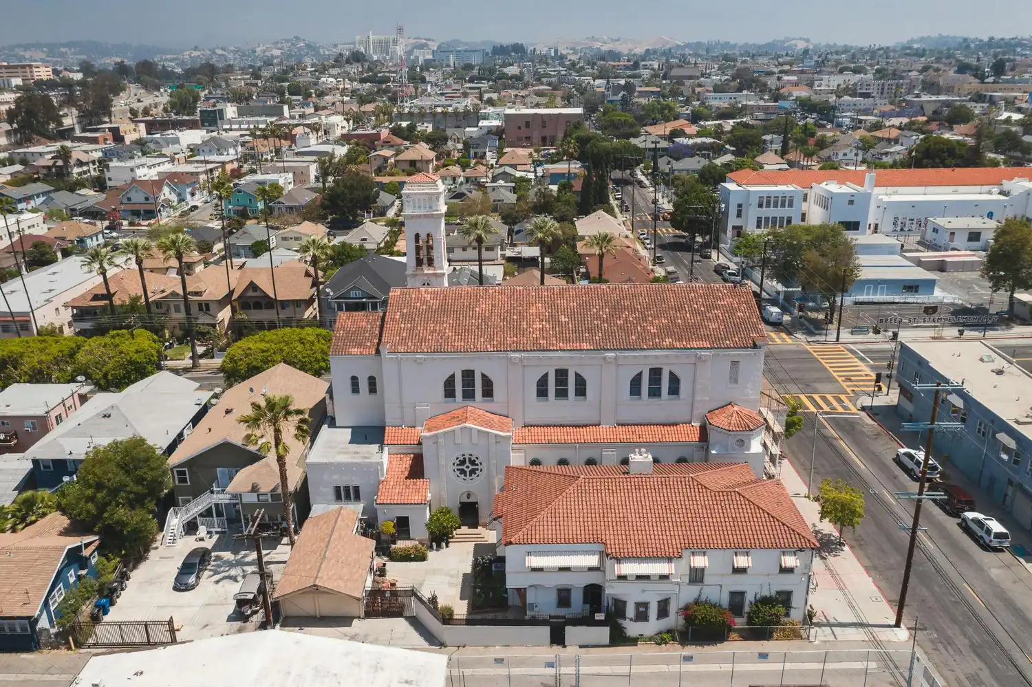 Aerial view of a church with red tile roofing surrounded by homes, streets, palm trees, and city buildings in the distance.