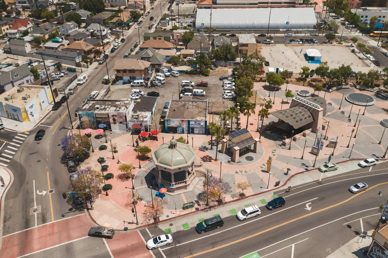 Aerial view of a downtown plaza with nearby streets, storefronts, parked cars, homes, and a small pavilion.