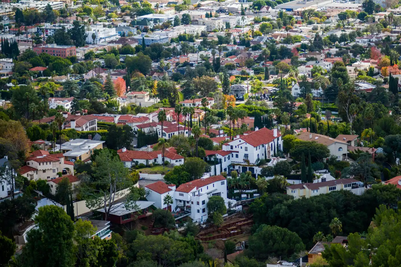 Aerial view of a hillside neighborhood with Spanish-style homes, red tile roofs, trees, and city buildings in the background.