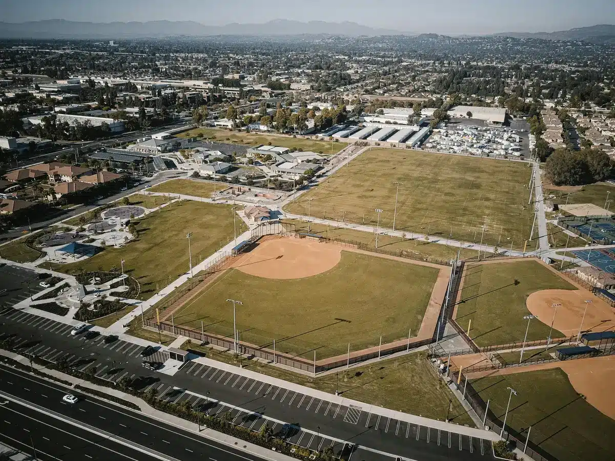 Aerial view of a large sports park with baseball fields, open grass areas, parking lots, nearby neighborhoods, and distant mountains.
