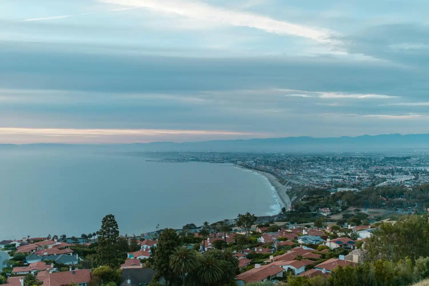 Coastal hillside view with homes, trees, ocean shoreline, a bay, distant city lights, and a cloudy evening sky.