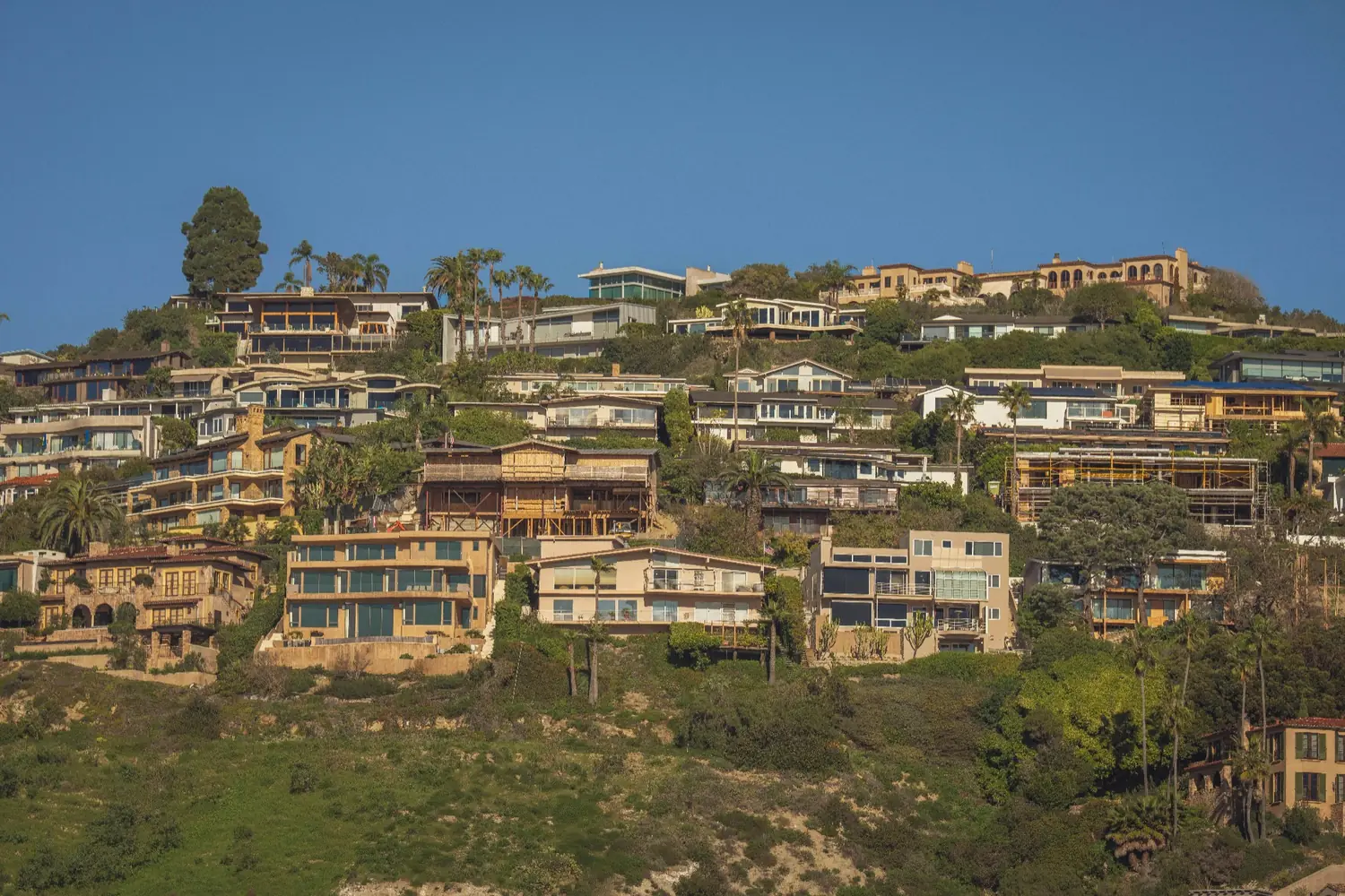 Hillside homes with modern and luxury architecture surrounded by trees, greenery, and a clear blue sky.