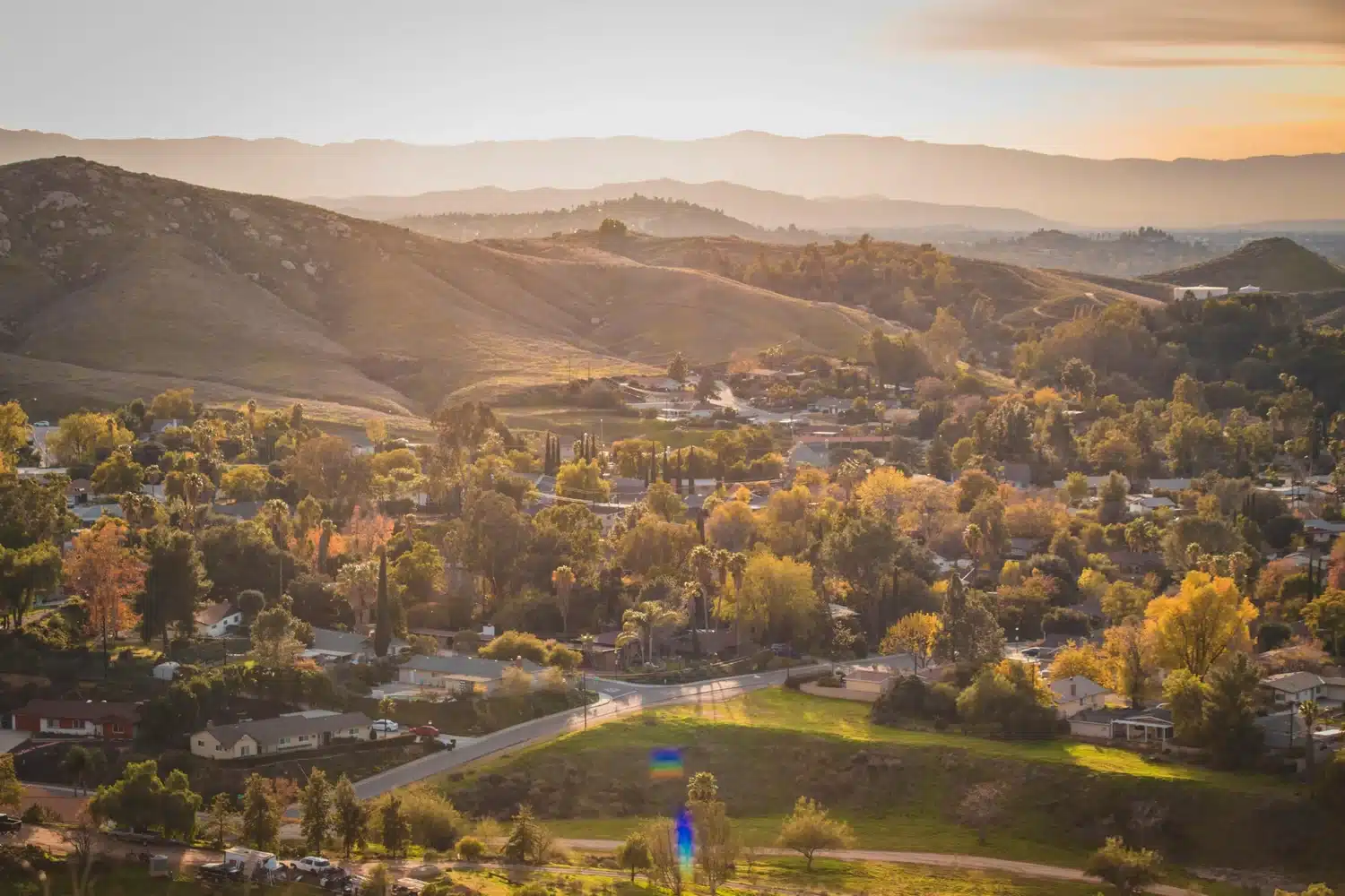 Scenic sunset view of a residential neighborhood surrounded by trees, rolling hills, and warm golden light.