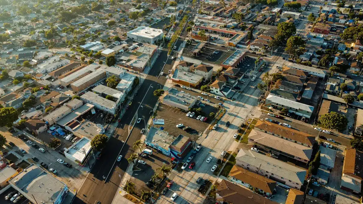 Aerial view of a city neighborhood with streets, homes, commercial buildings, parked cars, and warm evening light.